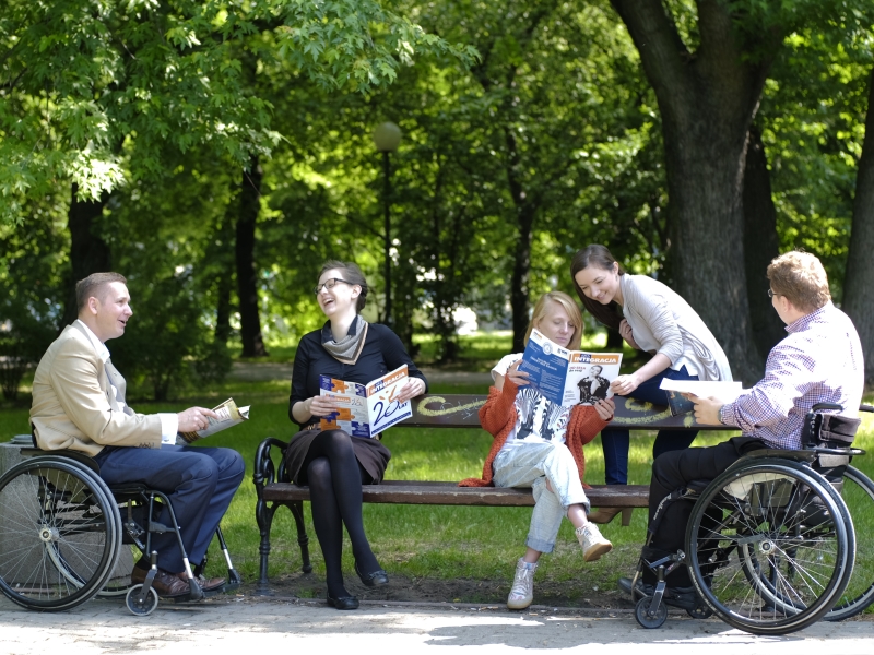 Osoby siedzące w parku i czytające magazyn Integracja, wsród nich dwóch mężczyzn poruszajacych sie na wózku/ fot. Piotr stanisławski