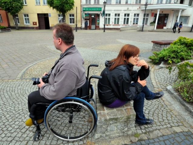 osoba na wózku i druga osoba siedzą do siebie tyłem na placu w jakimś mieście