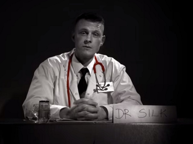 Raper ze złożonymi rękoma siedzi za biurkiem, ubrany w strój lekarza. obraz jest czarnobiały, oprócz czerwonego st etoskopu