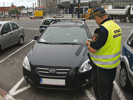 Strażnik miejski wypisuje mandat, stojąc przy samochodzie zaparkowanym na kopercie