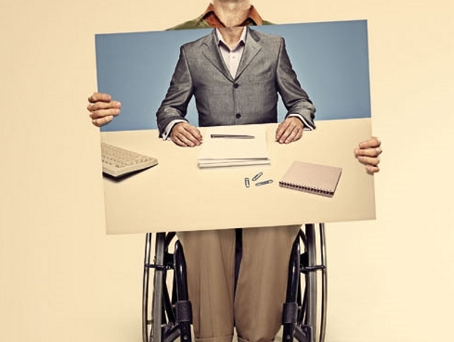 mężczyzna na wózku zwyczajnie ubrany trzyma tabliczkę ze swoim zdjęciem w garniturze przy pracy z komputerem 
