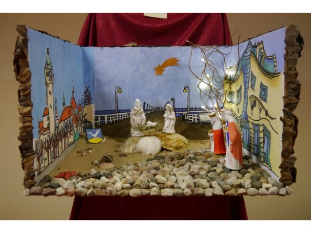bożonarodzeniowa szopka, gdzie Józef, Maryja i Jezus są na piasku, obok nich muszelki, za nimi molo sopockie, z prawej strony Krzywy Dom, a z lewej sopocka latarnia morska