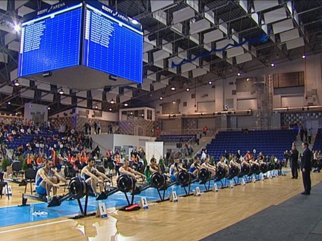 zawodnicy siedzący na ergometrach w jednym rzędzie oczekują na start rundy. W tle widać krzesełka kibiców na hali, w której odbywały się zawody. Nad głowami sportowców jest ekran informujący o wynikach