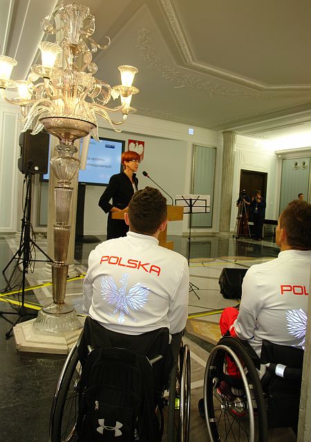 Minister Elżbieta Rafalska przemawia podczas konferencji. Na pierwszym planie paraolimpijczycy na wózkach