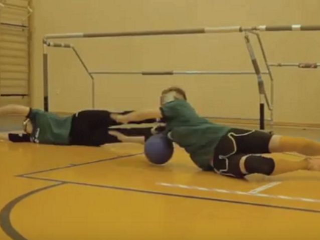 dwóch zawodników broni bramki przed piłką leżąc na podłodze i zasłaniając ciałem bramkę