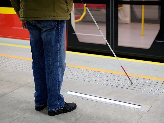 Niewidoma osoba z białą laską stoi przed wejściem do wagonu metra