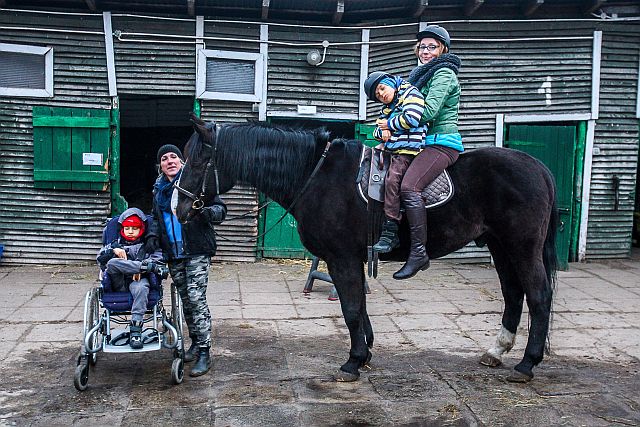 Przed stajnią. Vanessa Grzybowska siedzi na koniu z jednym ze swoich dzieci, obok stoi opiekunka z drugim dzieckiem na wózku