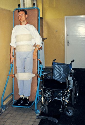 Piotr Pawłowski przypięty do pionizatora, obok stoi wózek inwalidzki