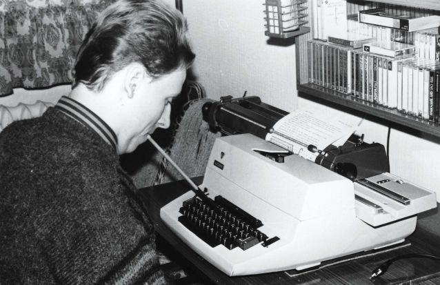 Czarno-białe zdjęcie Piotra Pawłowskiego piszącego na maszynie trzymanym w ustach patyczkiem