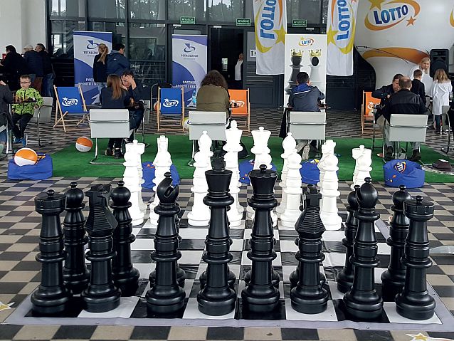 Ogromna stojąca na podłodze szachownica z figurami, w tle grający szachiści