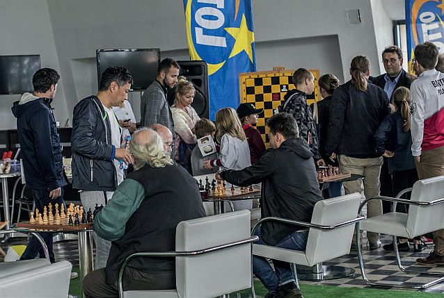 Kilku mężczyzn gra w szachy, w tle chodzą widzowie