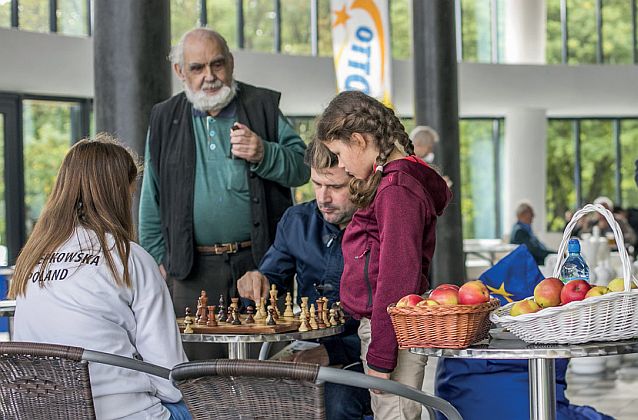 Kobieta i mężczyzna grają w szachy, przyglądają się im dziewczynka i starszy siwy pan
