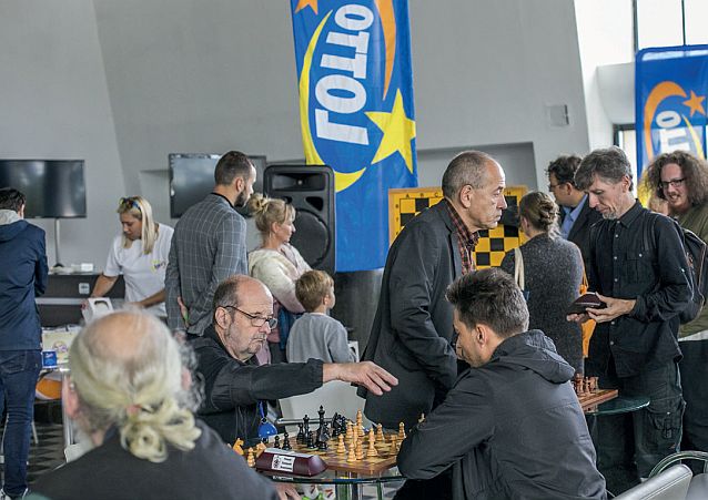 Dwaj mężczyźni grają w szachy, wokół chodzą widzowie