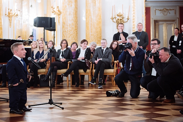 Andrzej Łukasik przemawia do mikrofonu w eleganckiej sali Zamku Królewskiego. Przed nim klęczą fotoreporterzy, w tle publiczność