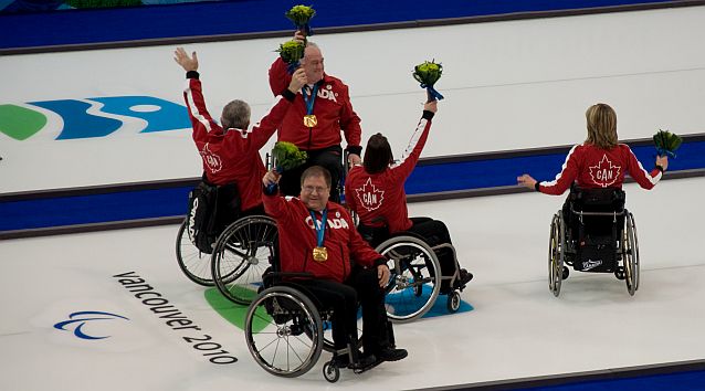 Zawodnicy na wózkach stoją na lodowym torze do curlingu z napisem Vancouver 2010. Mają medale na szyjach, cieszą się, machają do publiczności
