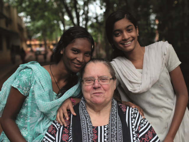 W środku siedzi Helena Pyz - kobieta ok. 50 lat, ma okrągły kształt twarzy, okulary i zaczesane do tyłu włosy. Obok niej po prawej i lewej znajdują się dwie młode dziewczyny z Indii. Wszystkie kobiety są spokojne i uśmiechnięte