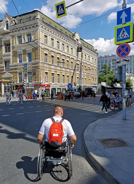 Mężczyzna na wózku na jezdni ulicy w Moskwie. W tle klasycystyczny gmach, obok przejście dla pieszych i przechodnie