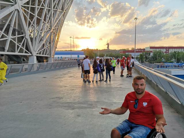 Łukasz Irla rozkłada ręce na tle stadionu i zachodzącego słońca, będąc na moście po przegranym przez Polskę meczu z Japonią.