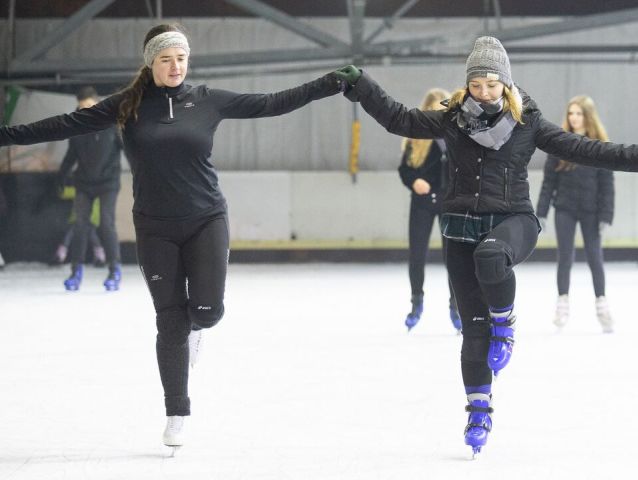 trenerka trzyma za dłoń dziewczynę, obie unoszą jedną nogę. są w łyżwach na lodowisku