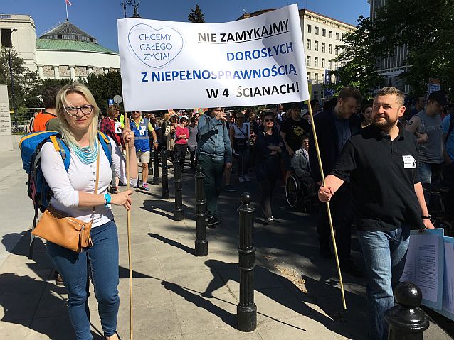 Manifestacja. Kobieta i mężczyzna niosą transparent z napisem: Chcemy całego życia. Nie zamykajmy dorosłych z niepełnosprawnością w 4 ścianach!
