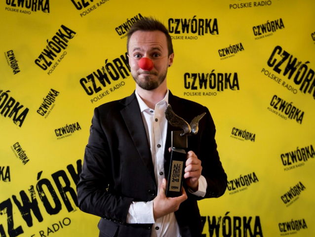 Michał Bańka ma na nosie czerwoną gąbkę, jaką mają klauni, w dłoni trzyma statuetkę Nieprzecietnego 2018 