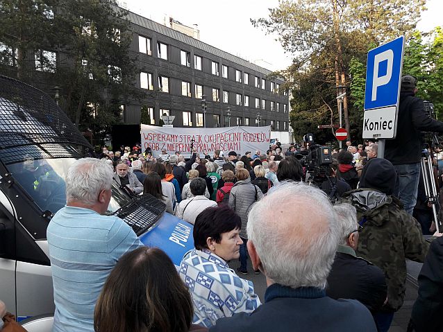 Manifestacja na ulicy, w oddali duży transparent z napisem: wspieramy strajk okupacyjny rodzin osób z niepełnosprawnościami