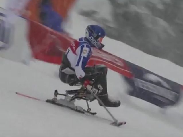 Jadący po śniegu zawodnik siedzący na sledżu z jedną nartą