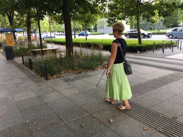 Kobieta w zielonej, długiej spódnicy idzie z opaską na oczach, trzymając białą laskę po ścieżce dla osób niewidomych