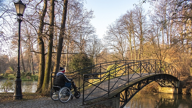 Mężczyzna na ręcznym wózku inwalidzkim z podpiętym z przodu napędem elektrycznym wjeżdża na mostek w parku