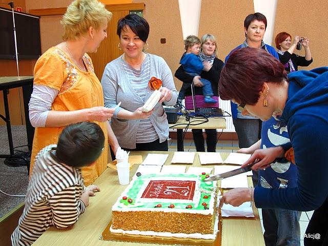 Na stole znajduje się tort z napisem na górze Zakątek 21. Tort kroi jedna z kobiet, obok stolika stoją dwie kobiety wraz z dzieckiem i rozmawiają uśmiechnięte