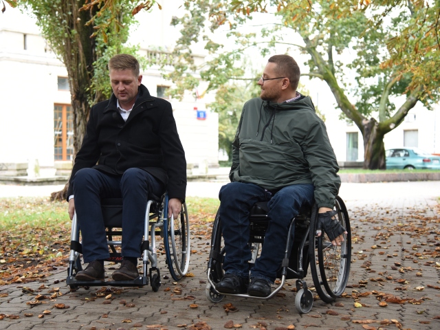 Andrzej Rozenek jedzie na wózku po chodniku w parku, obok niego mężczyzna na wózku. Rozmawiają