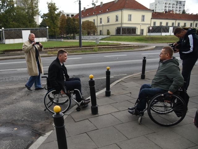 Andrzej Rozenek wjeżdża wózkiem na chodnik, starając się pokonać krawężnik. Na chodniku czeka na niego mężczyzna poruszający się na wózku