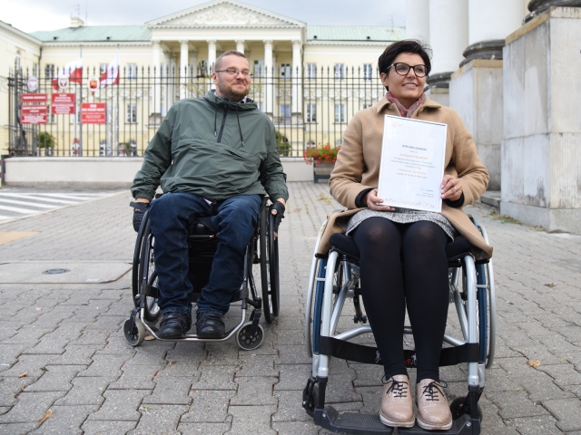 Justyna Glusman siedzi na wózku i trzyma w dłoniach dyplom. Delikatnie za nią znajduje się uśmiechnięty mężczyzna, poruszający się na wózku. Stoją przed bramą ratusza