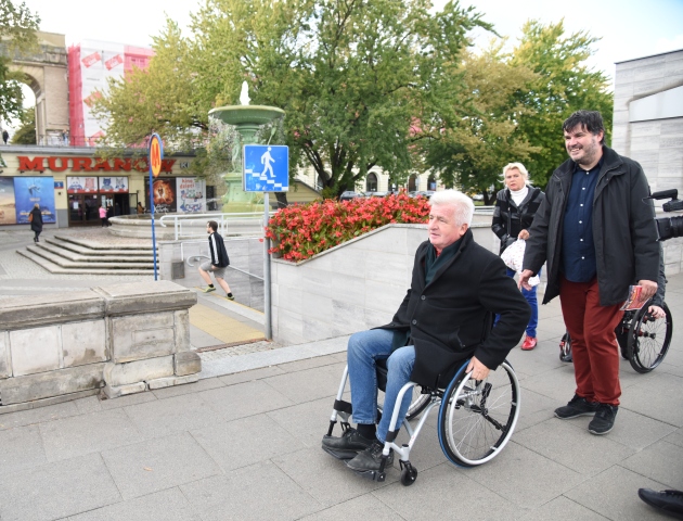 Piotr Ikonowicz porusza się na wózku po chodniku przy Kinie Muranów, tuż za nim idzie asekurant