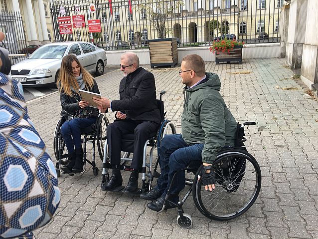 Sławomir Antonik na wózku przed bramą ratusza. Obok niego kobieta i mężczyzna - także na wózkach