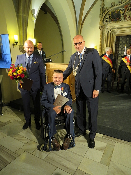 Bartłomiej Skrzyński z przypiętym medalem siedzi na wózku, nad nim stoją dwaj mężczyźni, w tym prezydent Wrocławia