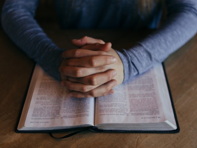 kobiece złączone dłonie jak do modlitwy leżą na Piśmie świętym, które znajduje się na biurku