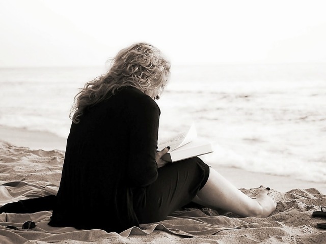kobieta siedzi bokiem na plaży, czyta książkę