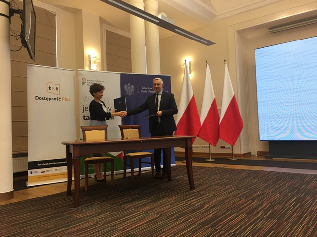 prezes PFRON maląg i minister Kuberski ściskają sobie ręce na tle 3 flag Polski