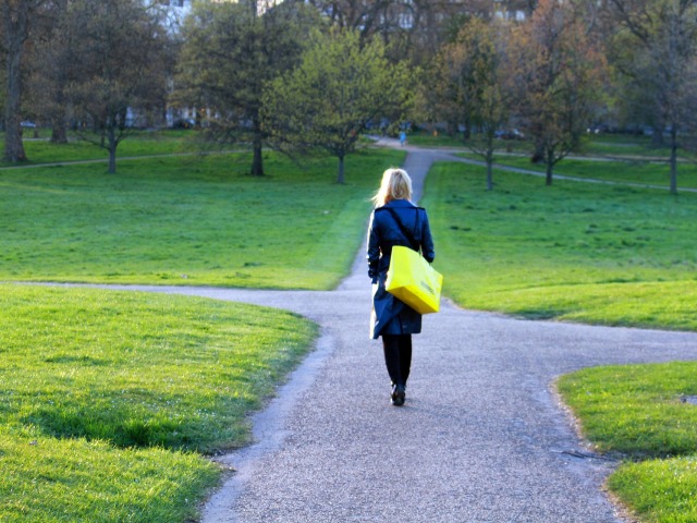 młoda kobieta w niebieskim płaszczu z żółtą torbą na ramieniu idzie parkiem widoczna z tyłu