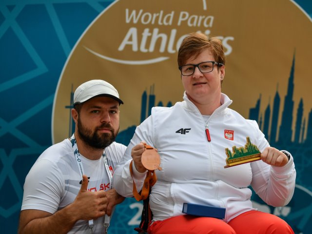 Lucyna Kornobys w jednej ręce trzyma medal w drugiej małą figurkę Dubaju obok mężczyzna z polskiej ekipy z kciukiem w górze