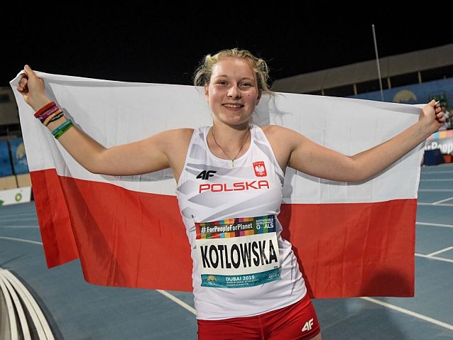 Faustyna Kotłowska w stroju reprezentacji Polski uśmiechnięta trzyma za plecami rozpostartą polską flagę