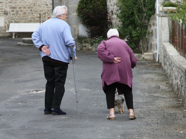 mężczyzna o kuli i kobieta zgarbiona starsza obok niego z psem idą ulicą tyłem do obiektywu