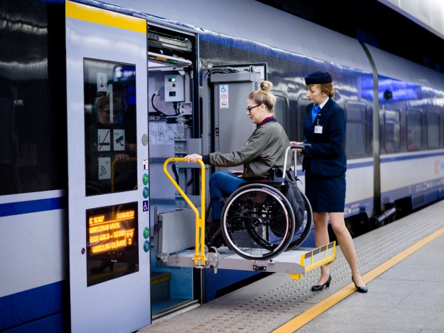 Pracownica PKP obsługuje rampę, na której znajduje się kobieta na wózku. Rampa przesuwa się z peronu w stronę wnętrza pociągu