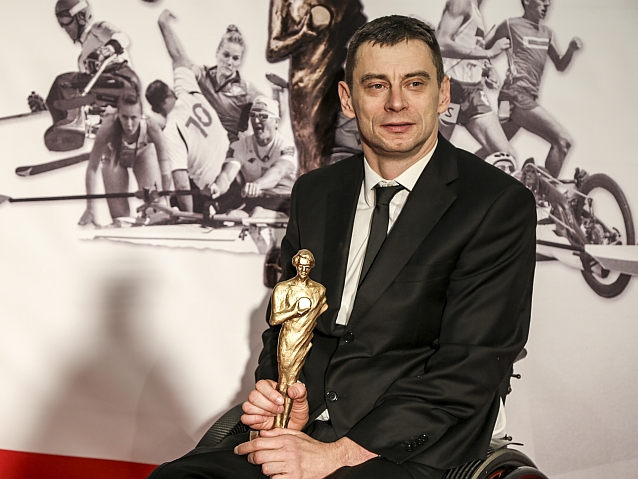 Jacek Czech siedzi trzymając statuetkę, w tle grafika przedstawiająca sportowców