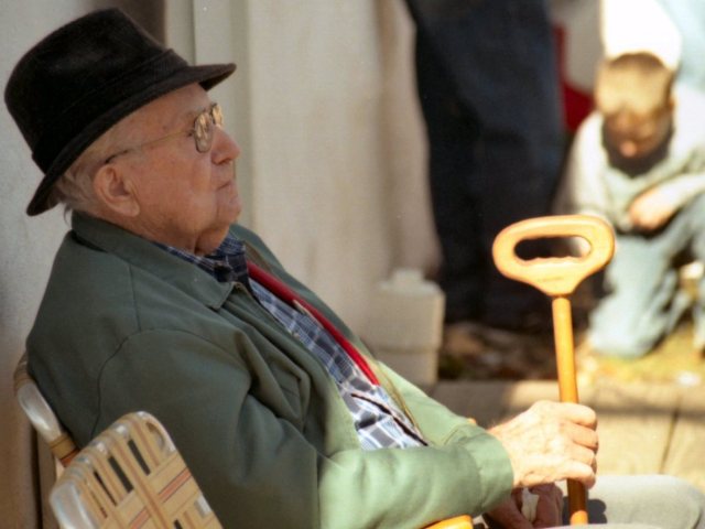 starszy pan w kapeluszu siedzi na ulicy na krześle opiera się na lasce