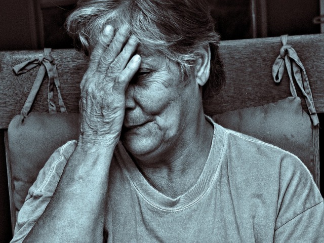 starsza kobieta siedzi na krześle, jedną dłonią przykrywa połowę twarzy