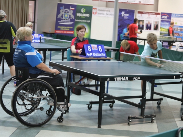 kobieta na wózku stoi przy stole do tenisa, w tle widać kobietę sędzię i chłopca ustawiającego piłkę na stole