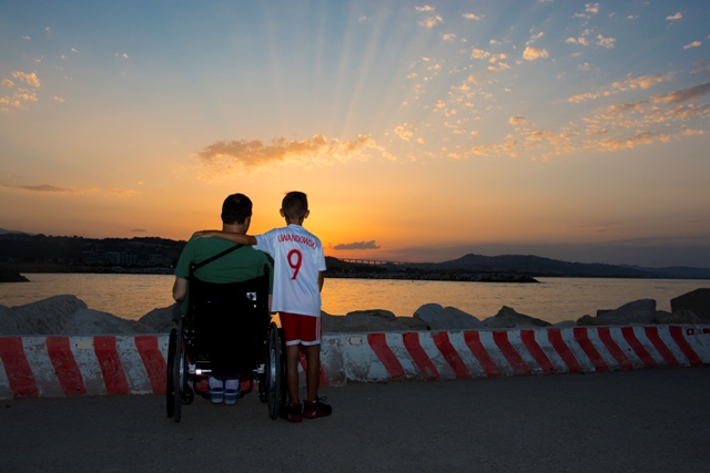 mężczyznę na wózku obejmuje chłopczyk. Stoją tyłem do odbiorcy, patrzą na zachodzące nad jeziorem słońce