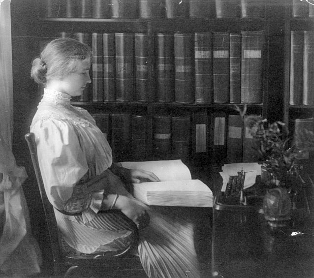 Helena Keller siedzi na krześle z książką napisaną w brajlu. W tle biblioteczka